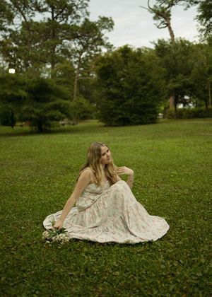 A person in a floral dress lies peacefully on a grassy lawn surrounded by trees on a sunny day.