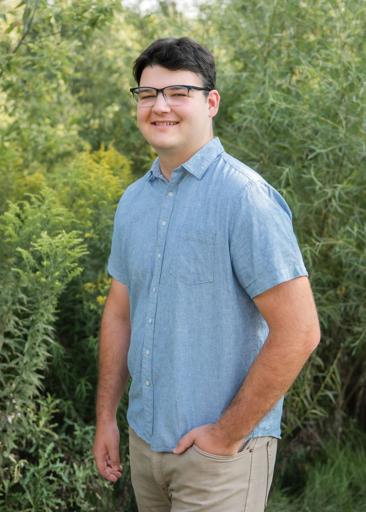 Someone wearing glasses and a light blue short-sleeve button down shirt poses outdoors against greenery in a casual portrait.