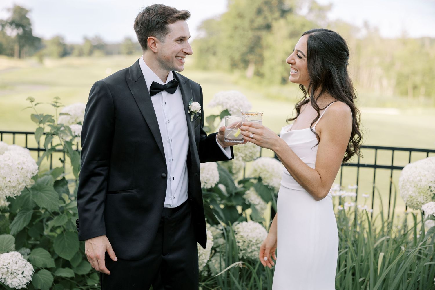 A joyful couple in formal wedding attire share a laugh together in an outdoor garden setting.