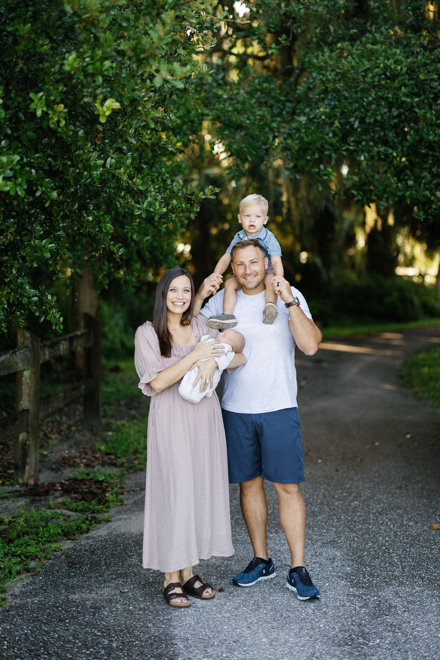 A couple stands with their baby on a tree-lined gravel path during an outdoor photo session.