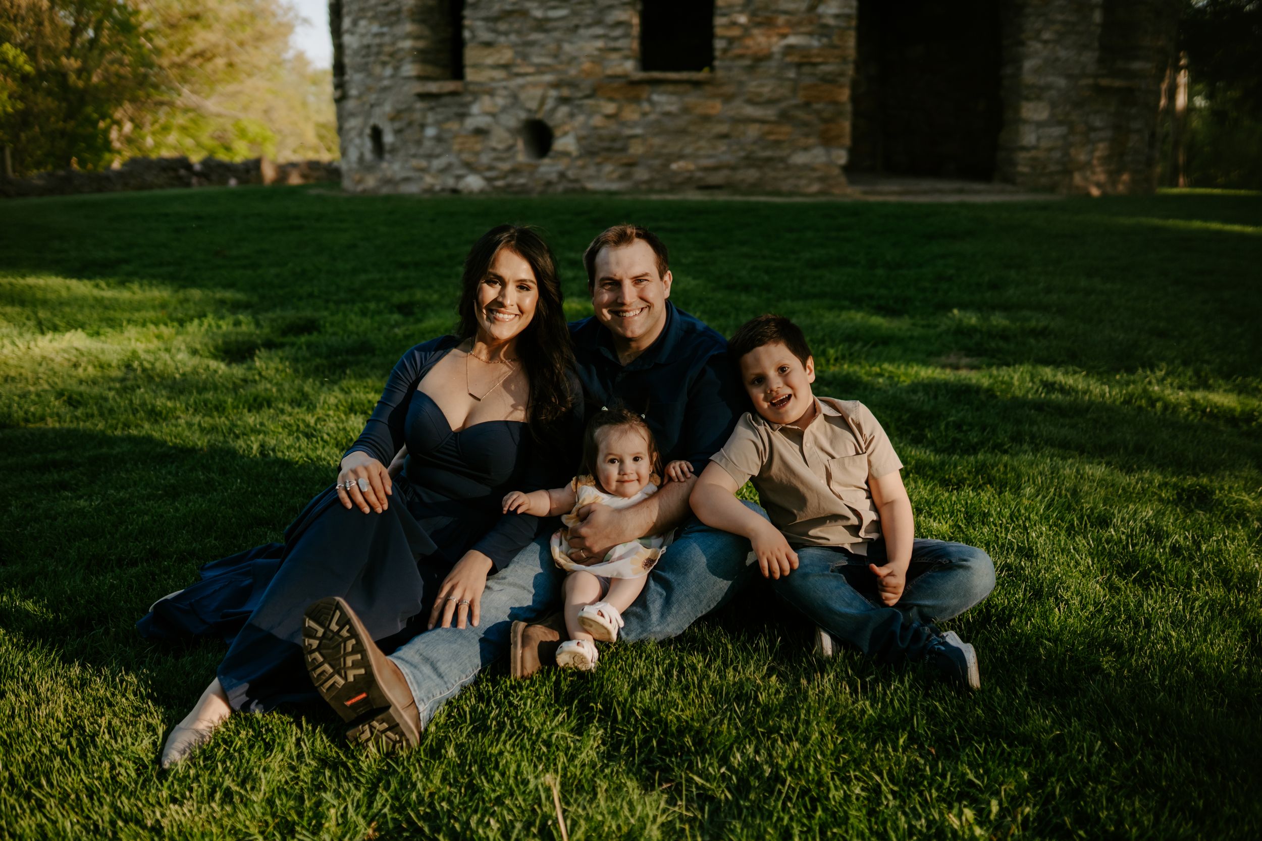 Family group relaxing together on grass near stone building at sunset.