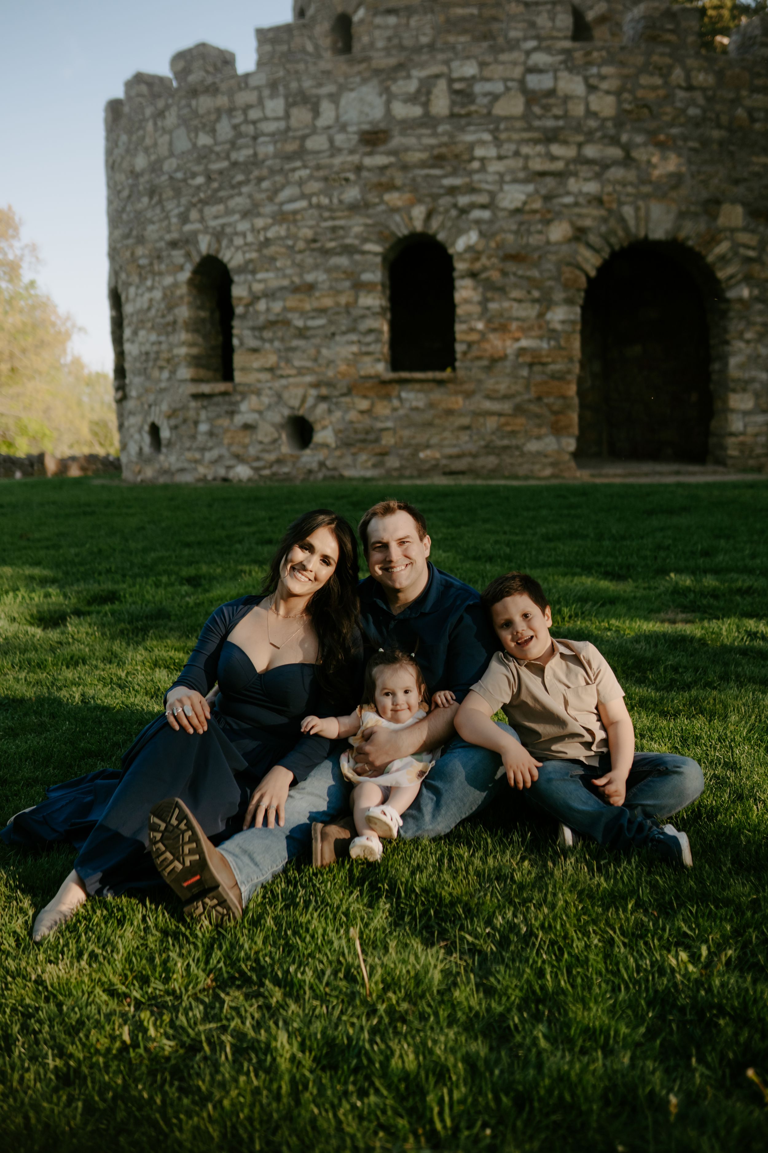 A family relaxes on green grass in front of a stone castle tower during a sunny afternoon photo session.