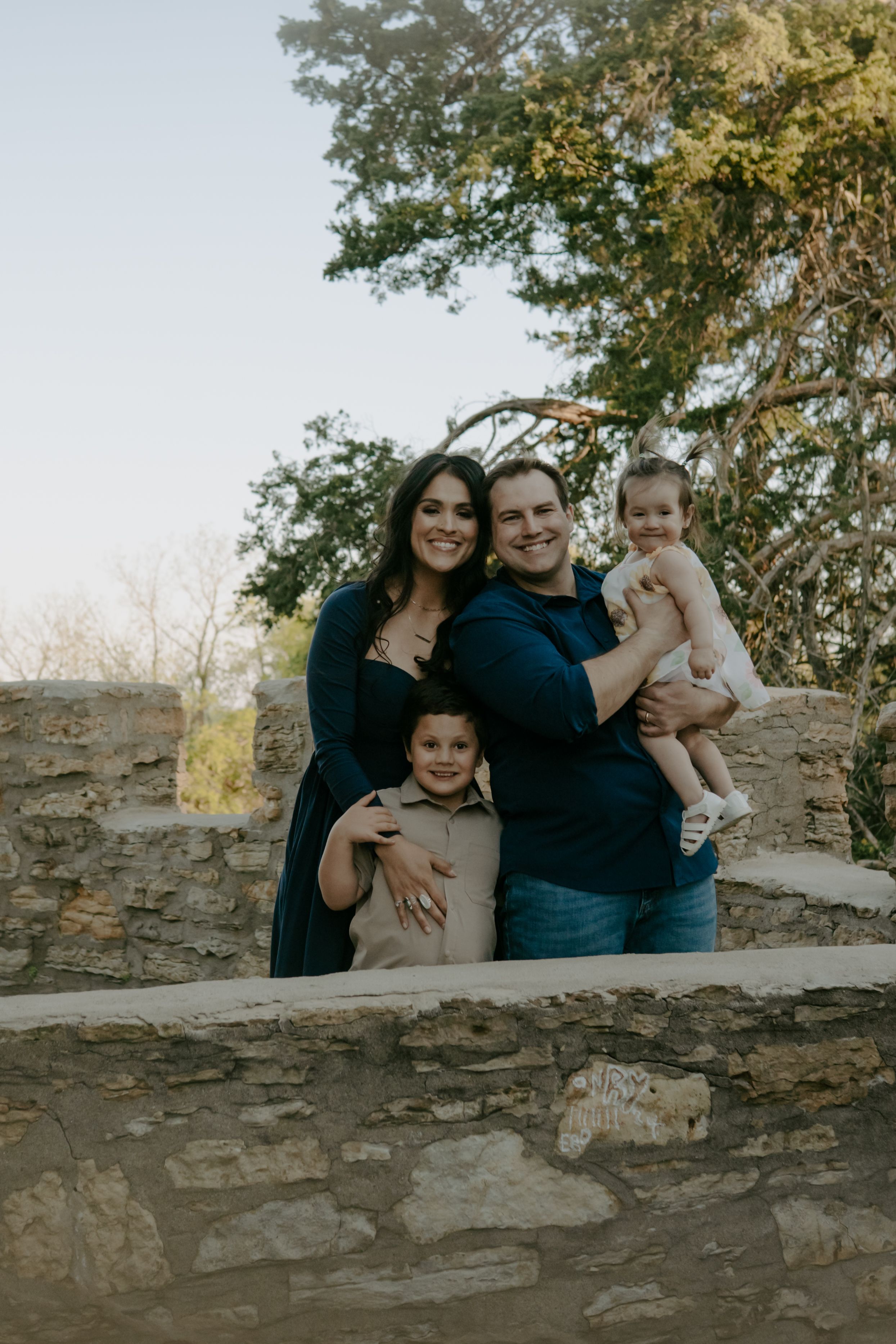 A family poses together against a stone wall with trees in the background during sunset.