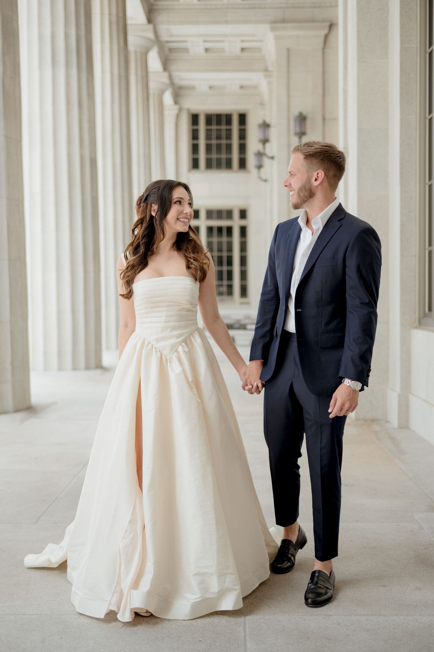 A couple in wedding attire share a joyful moment while walking together in an elegant columned hallway.