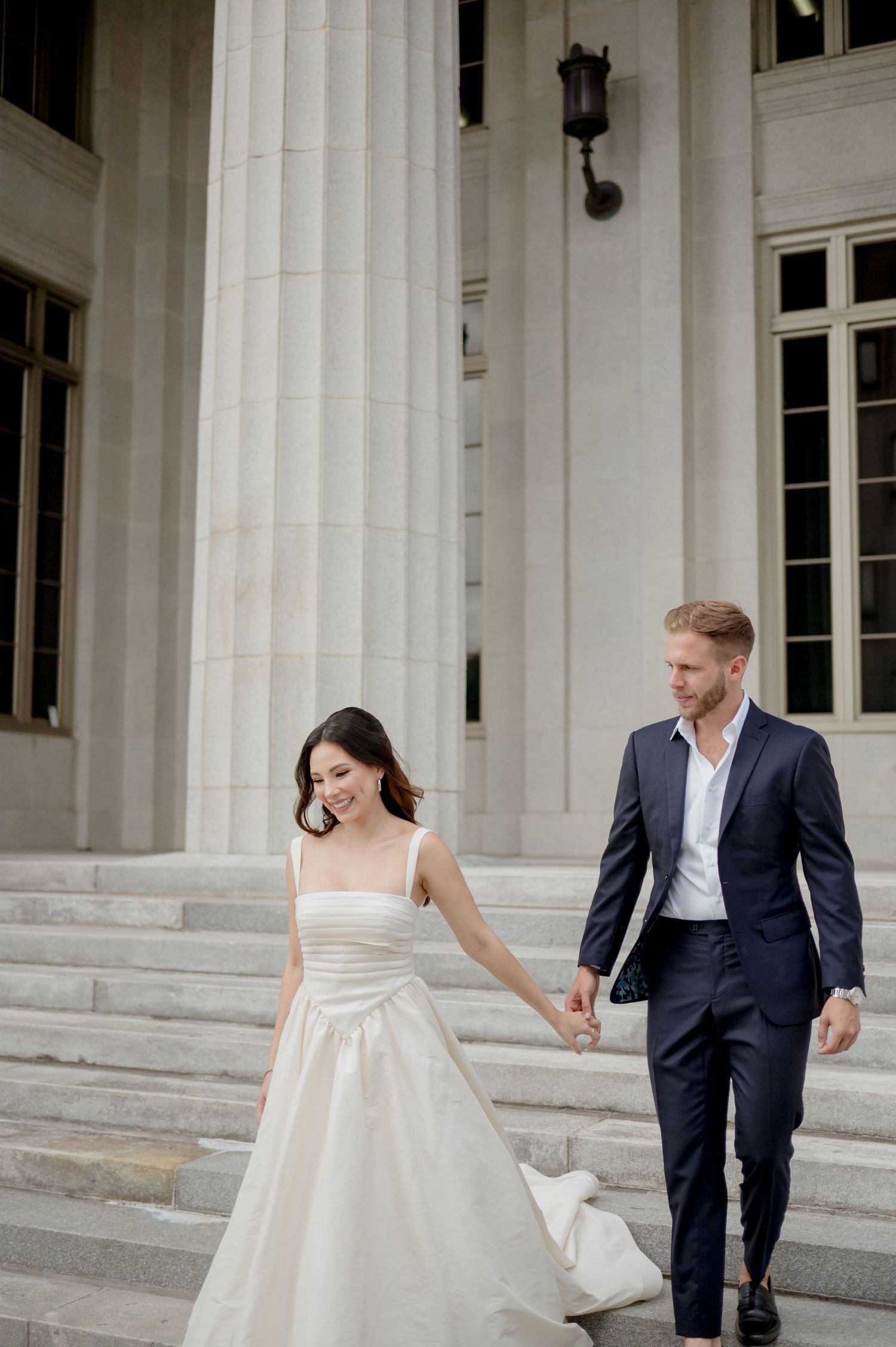 Elegant couple descends white stone stairs, bride in flowing ivory gown and groom in navy suit holding hands.