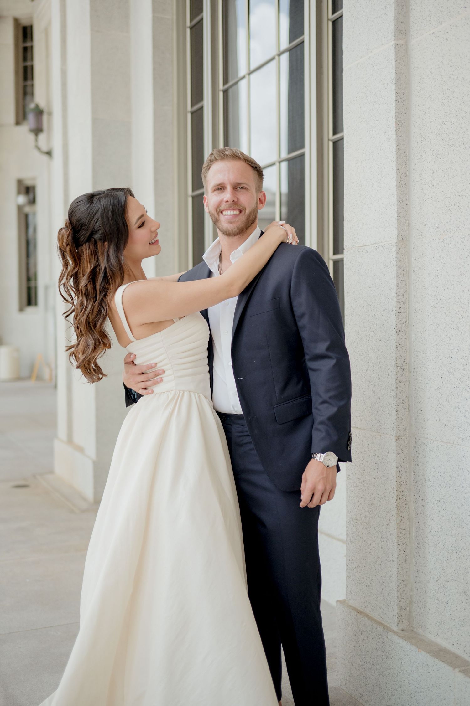A couple embraces and shares playful moments during a wedding photoshoot in an elegant white colonnade hallway.