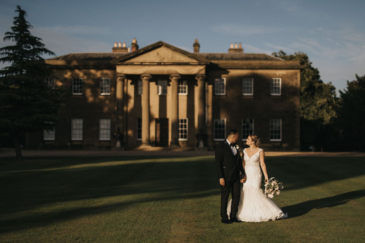 A couple poses on the manicured lawn in front of a grand columned mansion at golden hour.