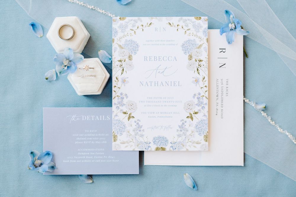 Wedding invitations in light blue and white with floral designs and gold accents arranged on a blue surface.