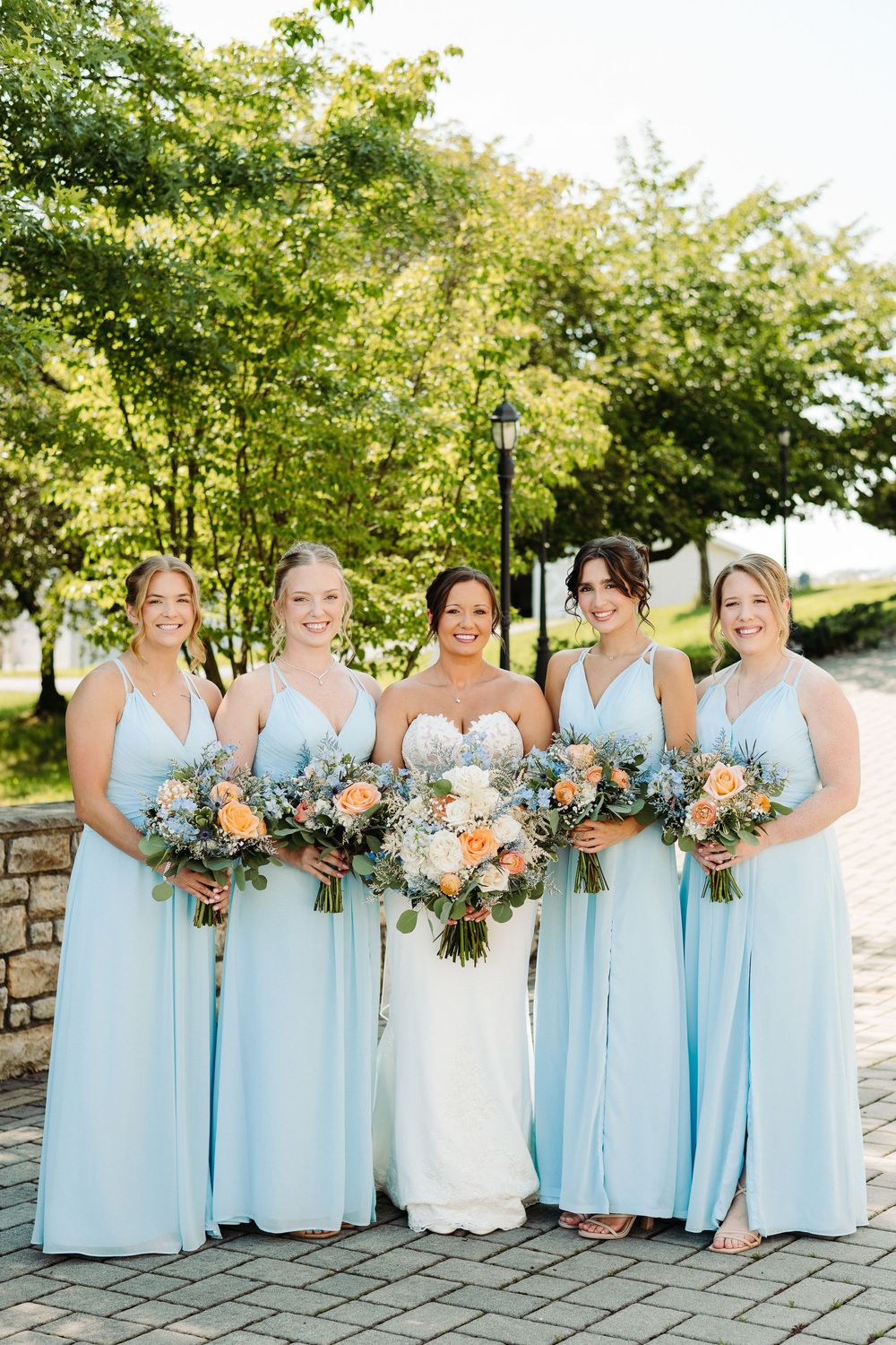 Wedding party in light blue bridesmaid dresses and white bridal gown pose together outdoors by stone wall.