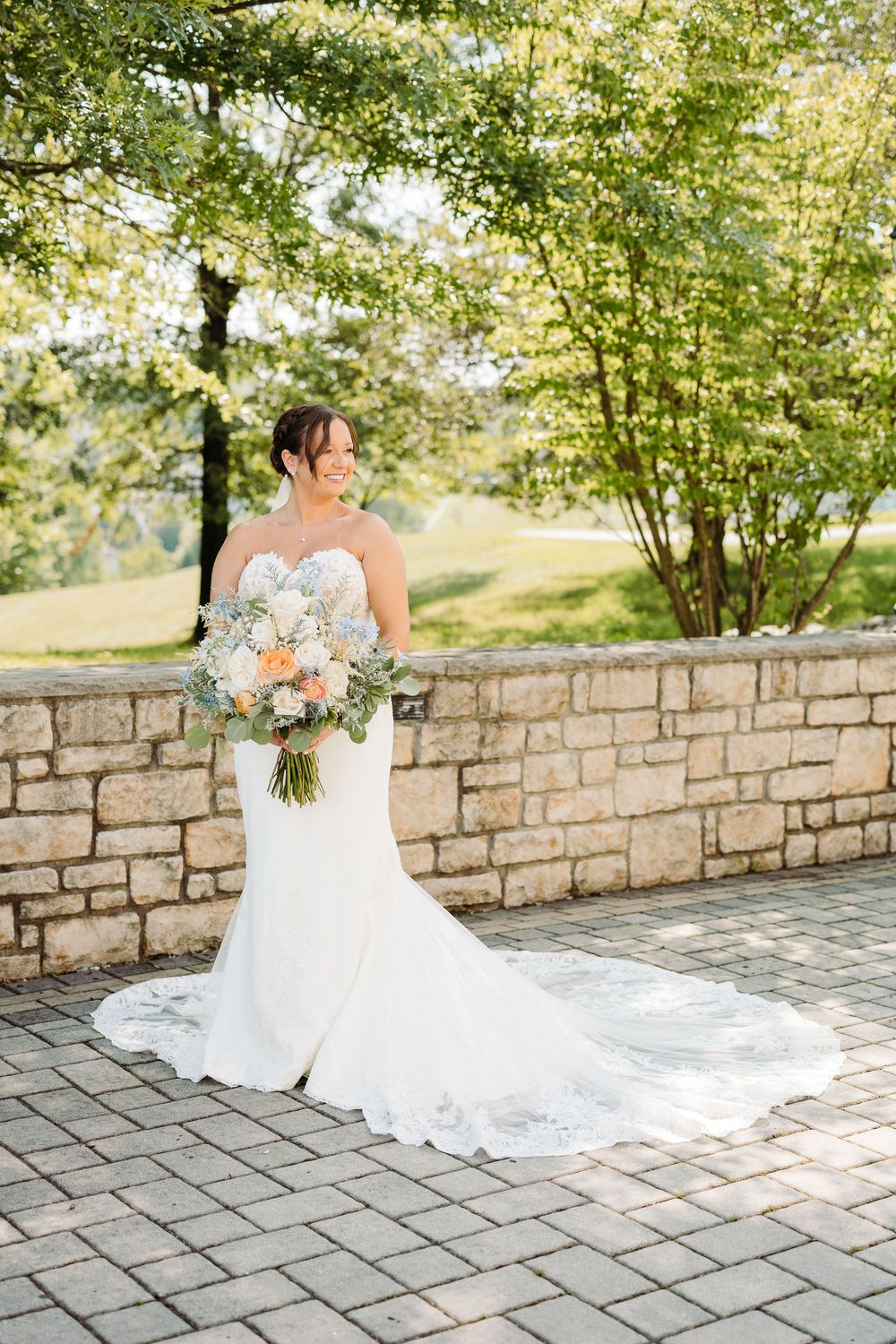 A wedding dress with delicate lace details flows elegantly against a stone wall backdrop in an outdoor garden setting.