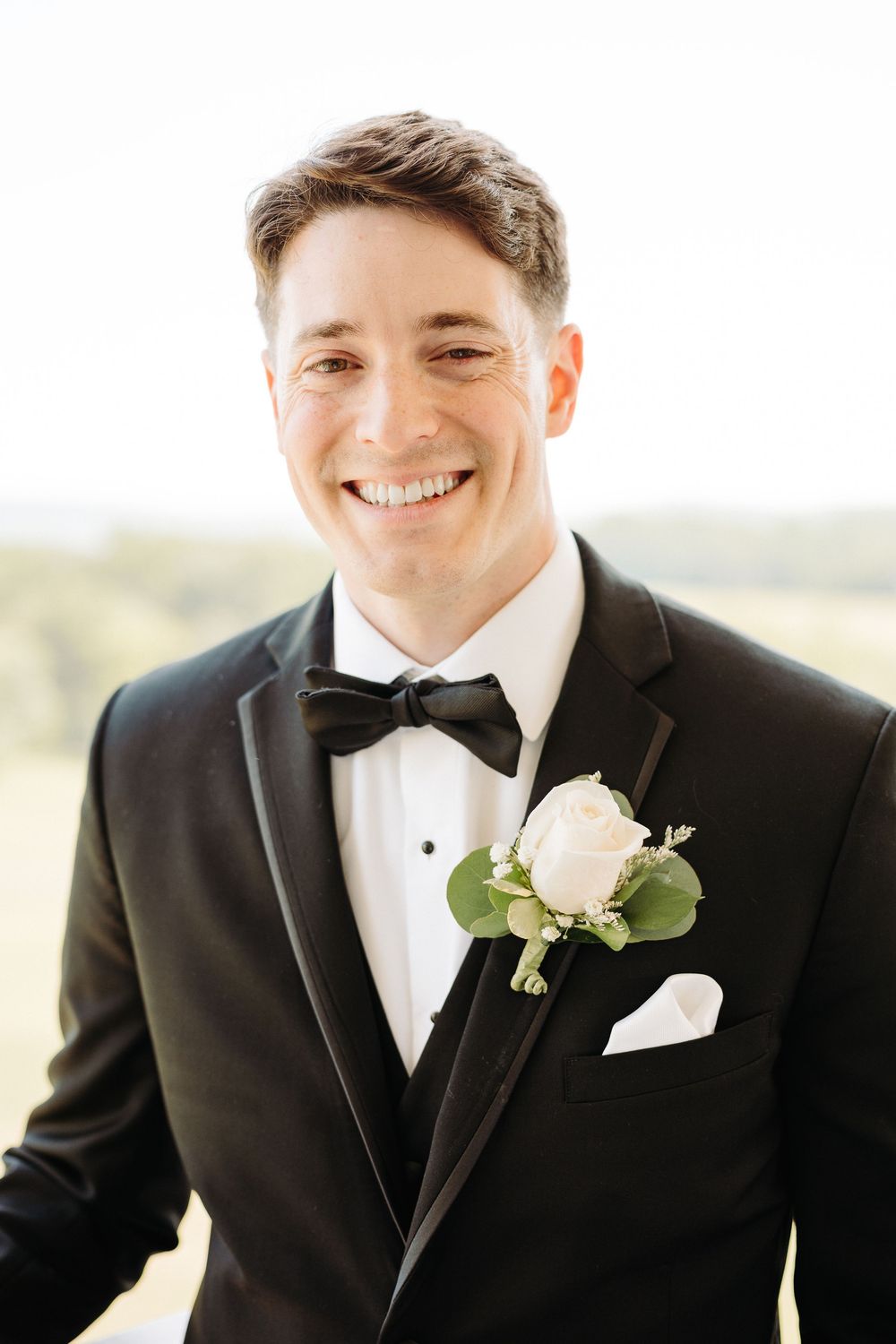 A groom wearing a classic black tuxedo with a white rose boutonniere smiles in outdoor portraits.