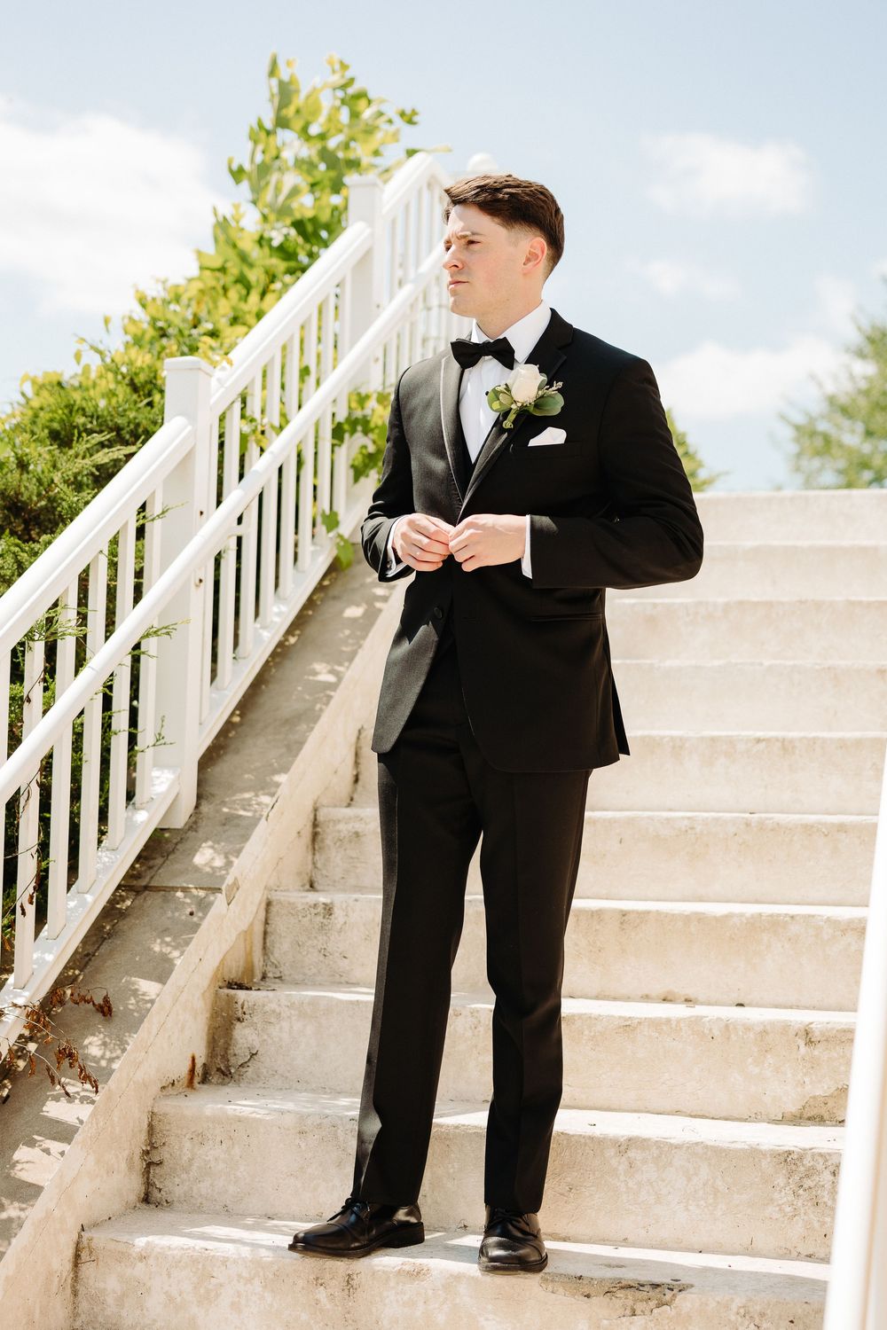 A person in a classic black tuxedo and bowtie stands on white stone stairs outdoors on a sunny day.