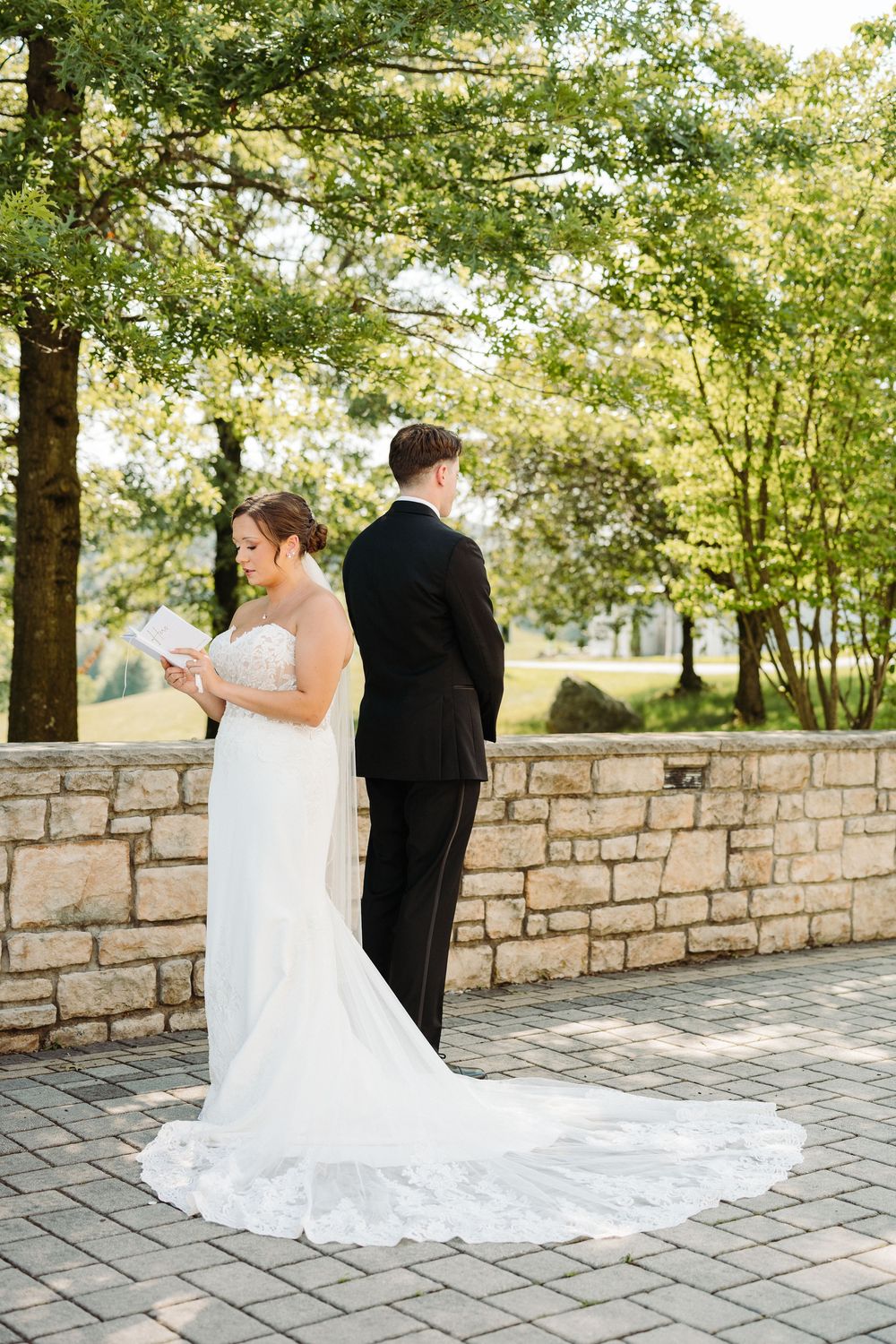 Bride in white gown and groom in black suit standing back-to-back at stone wall.
