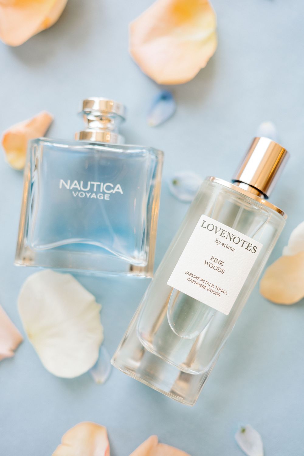 Two luxury perfume bottles of Nautica Voyage and Lovenotes fragrances surrounded by soft peach rose petals on blue surface.