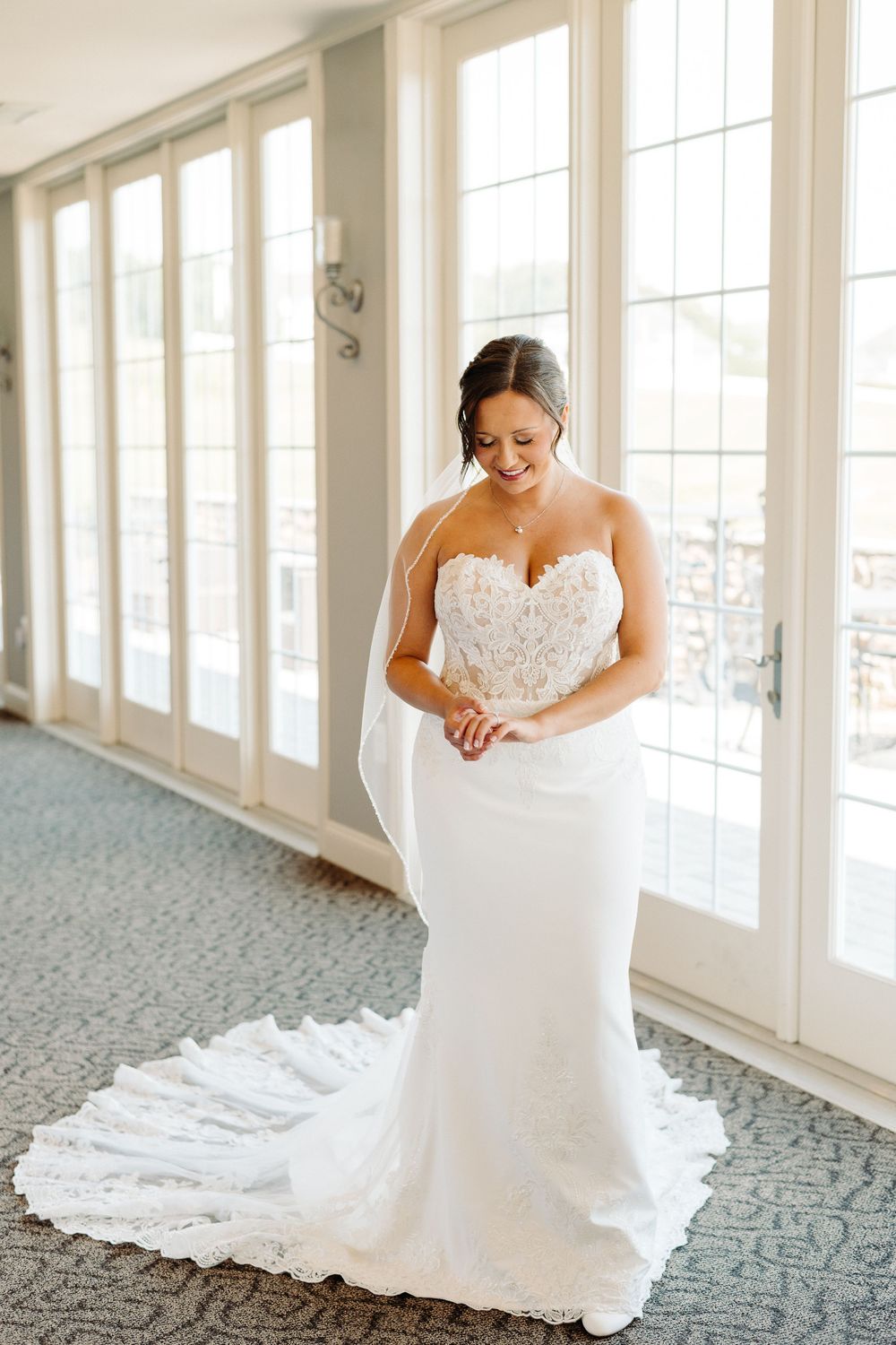 A bride in a strapless white wedding gown with lace details poses gracefully in a bright sunlit room.