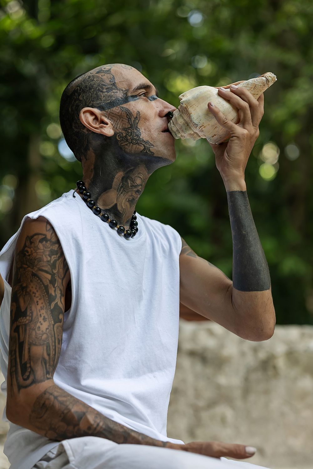 Person wearing white tank top with visible tattoos takes drink from glass bottle against blurred green background.