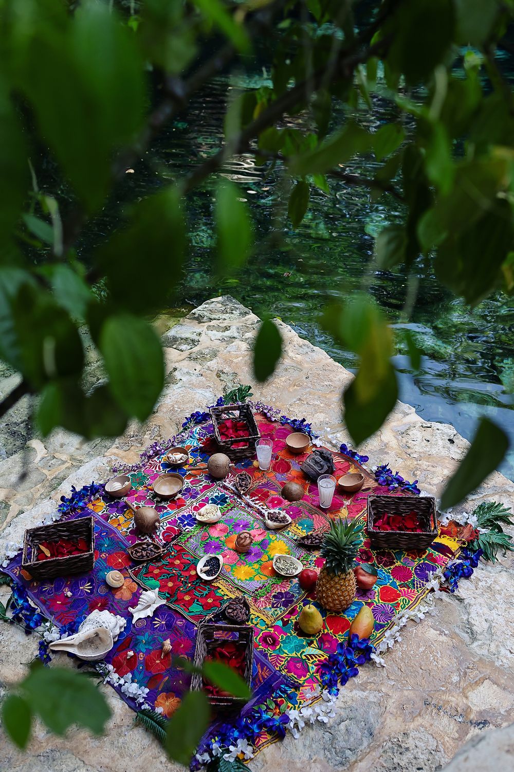 Colorful picnic blanket with red and blue textiles displays food and drinks by a tranquil pond framed by green leaves.