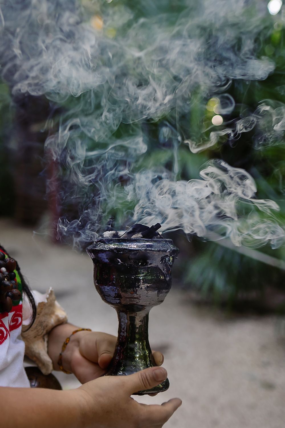 Smoke rises from a traditional ceramic incense burner being held against a blurred green background outdoors.