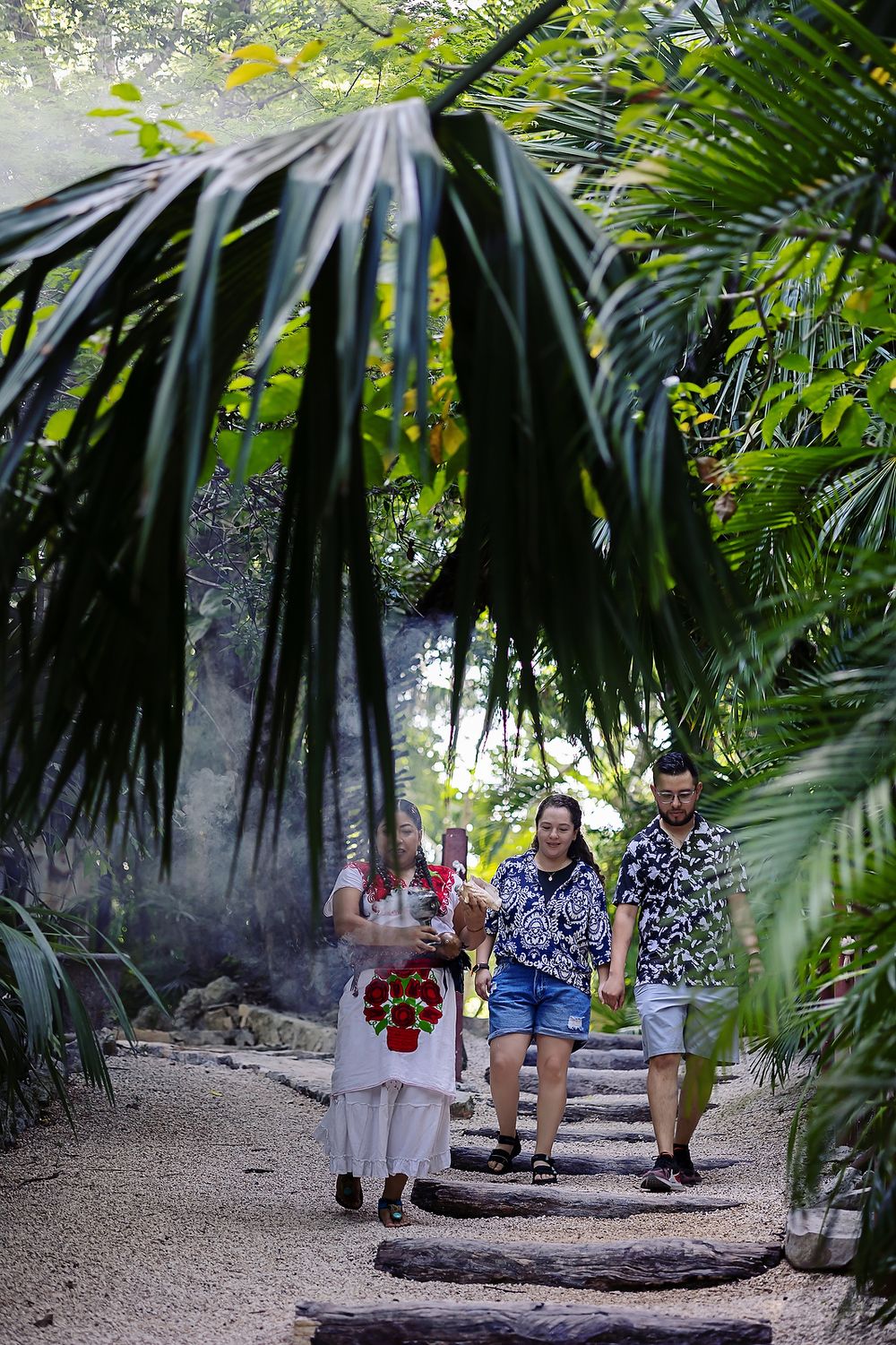 Visitors walking on a shaded tropical garden path lined with palm fronds and lush vegetation.