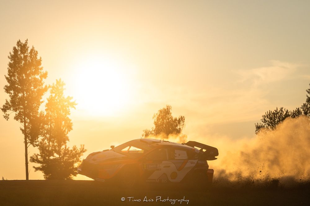 Rally car kicks up dust while racing against a dramatic golden sunset silhouetted by trees.