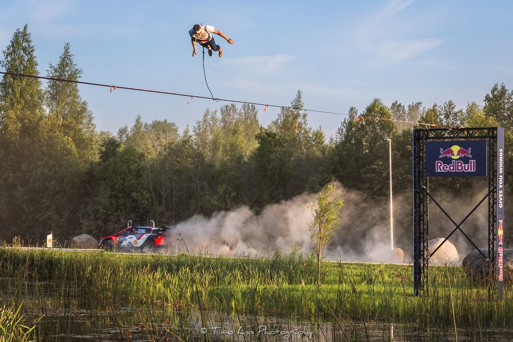 A person flying on a zipline above a dusty drift car track with trees in the background and a Red Bull sign.