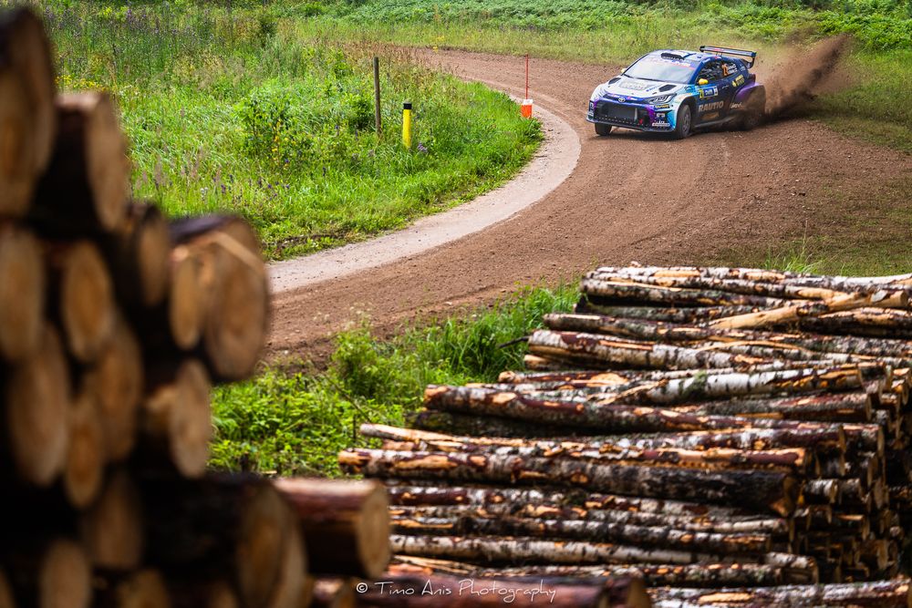 Rally car navigates dirt track corner past stack of timber logs during forest race stage.