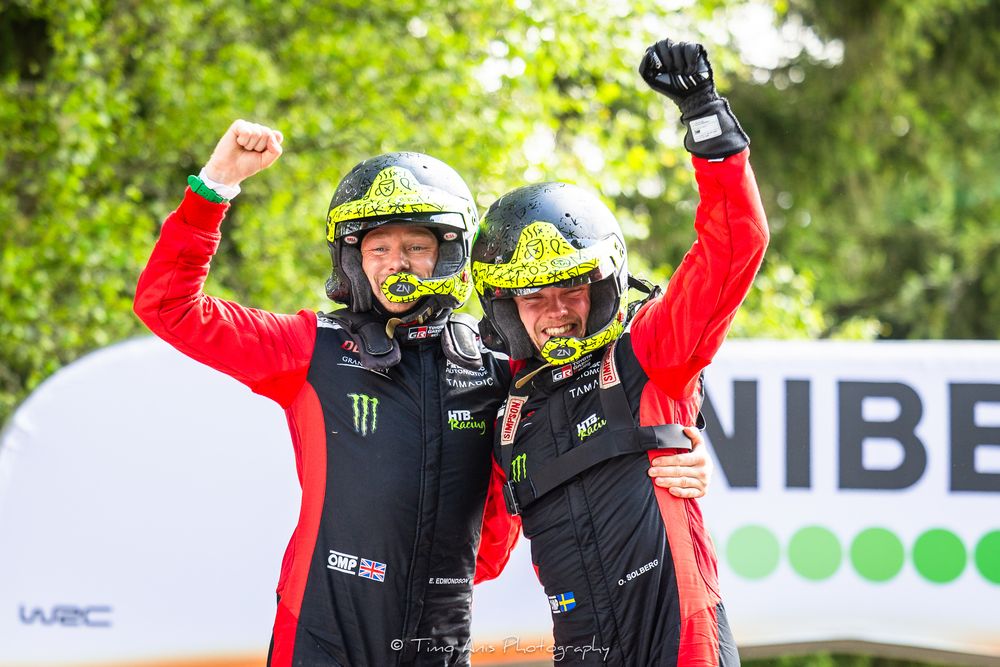 Rally drivers in red uniforms and helmets celebrate victory with raised arms at WRC racing event.