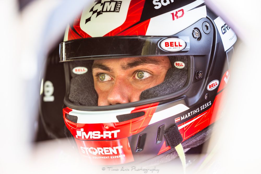 Close-up view of a race car driver wearing a red and black Bell racing helmet with MS-RT and SCM branding.