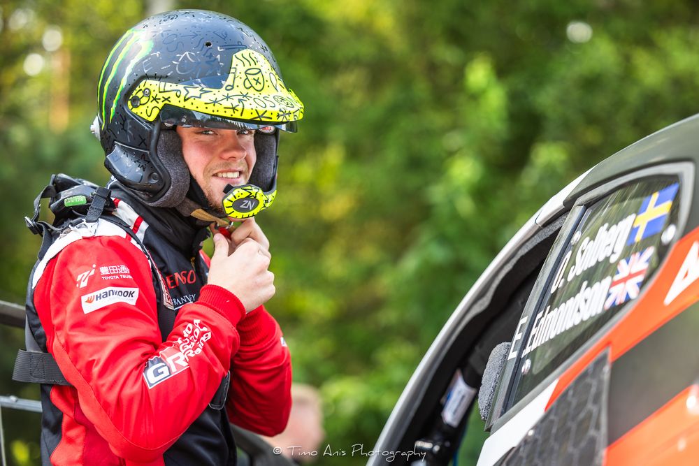Race car driver in red suit and black helmet adjusts equipment before competition at motorsport event.