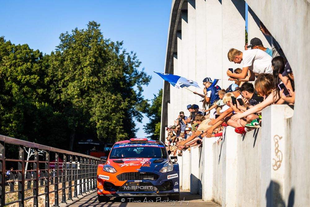Rally car races past excited spectators leaning over wall at motorsport event with trees in background.