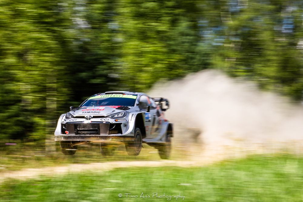 Rally car kicks up dust while drifting through a forest course with sunlight filtering through trees.