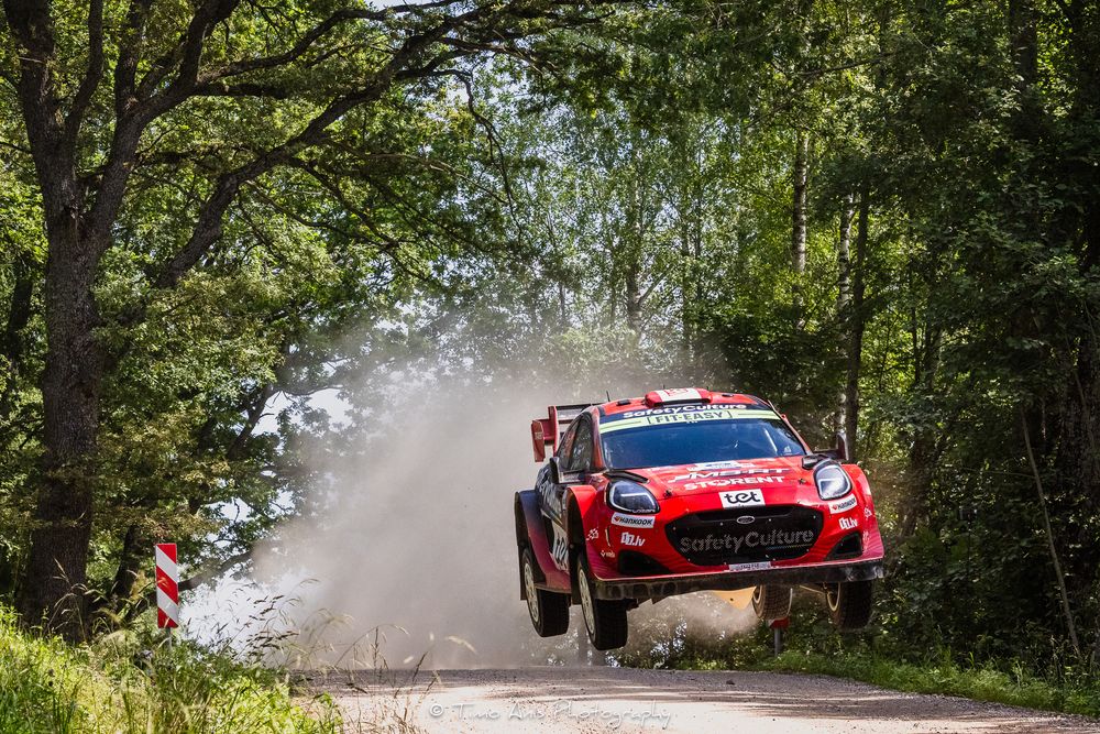 Red rally car catches air while jumping over a forest road during intense competition.