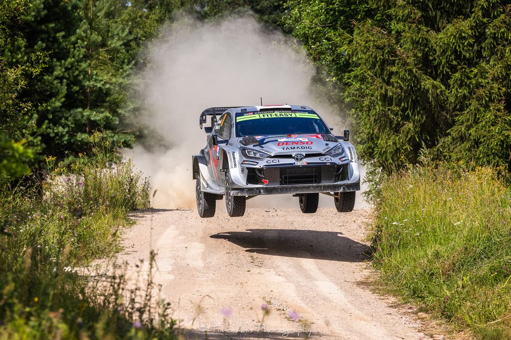 A rally car jumps over a dirt road during a race as dust kicks up behind it on a sunny summer day.