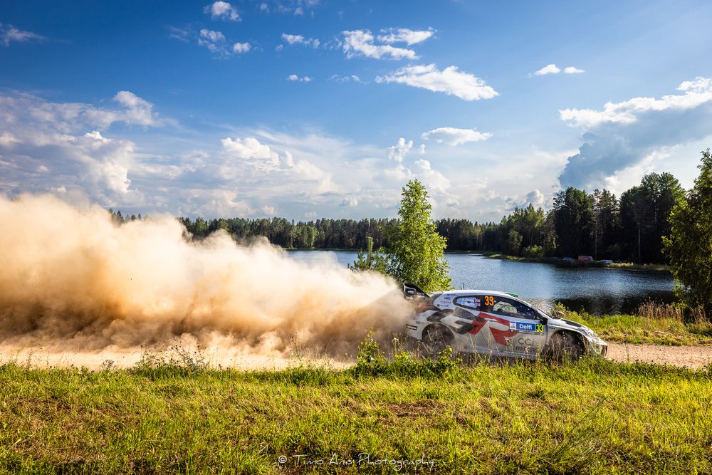 Rally car kicks up dust cloud while racing on grassy track beside a scenic lake under blue summer sky.