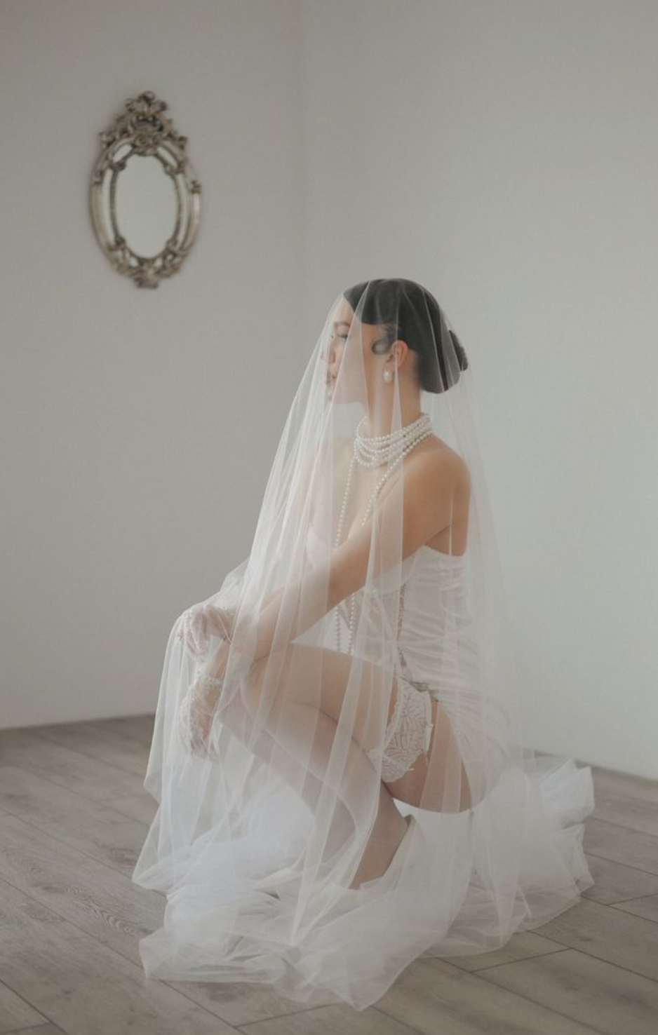 A bride in a flowing white wedding dress and veil poses gracefully in a minimalist room with a vintage mirror on the wall.
