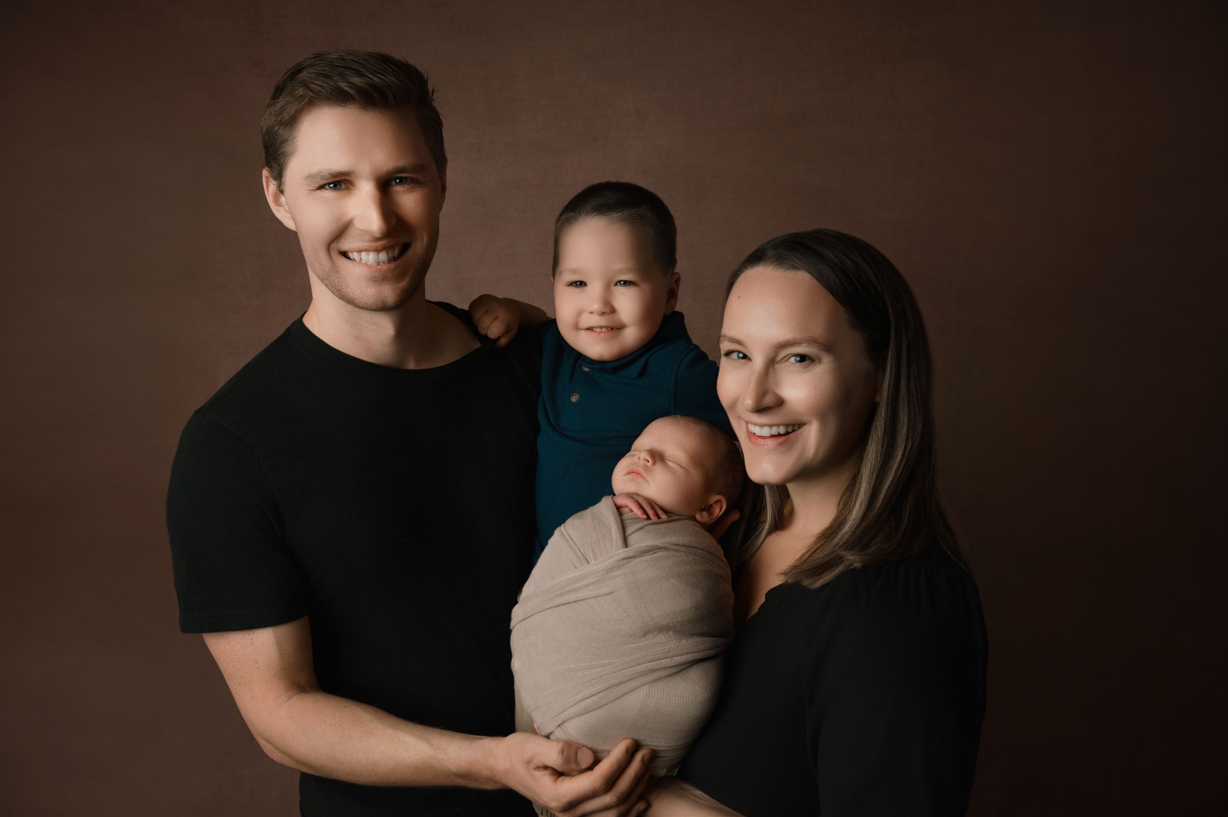 Family of four poses together against brown backdrop in studio portrait wearing black tops in warm lighting.