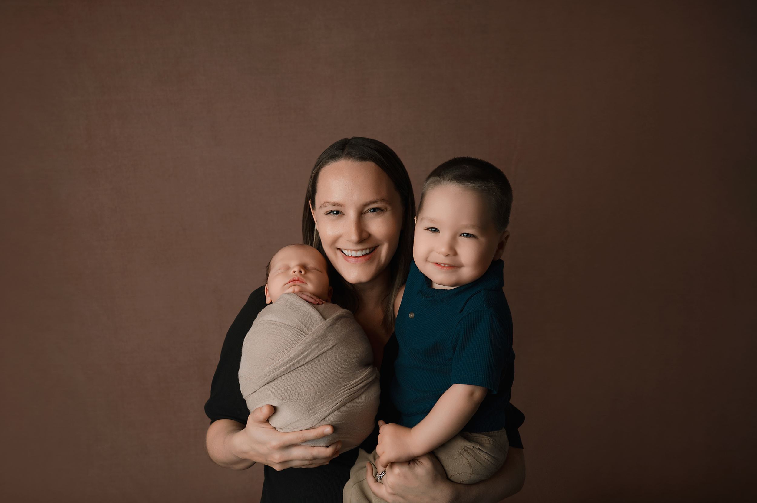 A heartwarming studio portrait against a brown background shows a parent with two young children, one in a dark shirt.