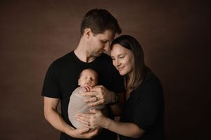 A couple in black shirts tenderly holds their newborn baby against a dark brown studio backdrop.