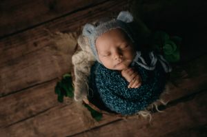 Sleeping newborn wrapped in a cozy blue swaddle rests on wooden floor with green foliage accents.