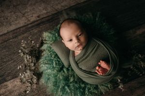Swaddled baby lying on textured wooden floor with emerald green tulle fabric and delicate white baby's breath flowers.