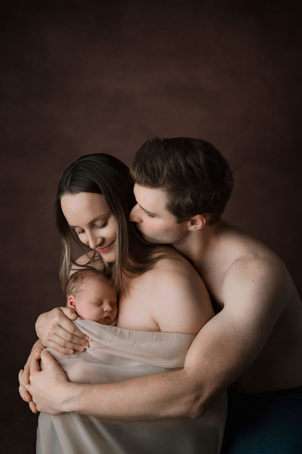 A tender intimate family portrait in soft warm lighting against a dark brown studio backdrop.