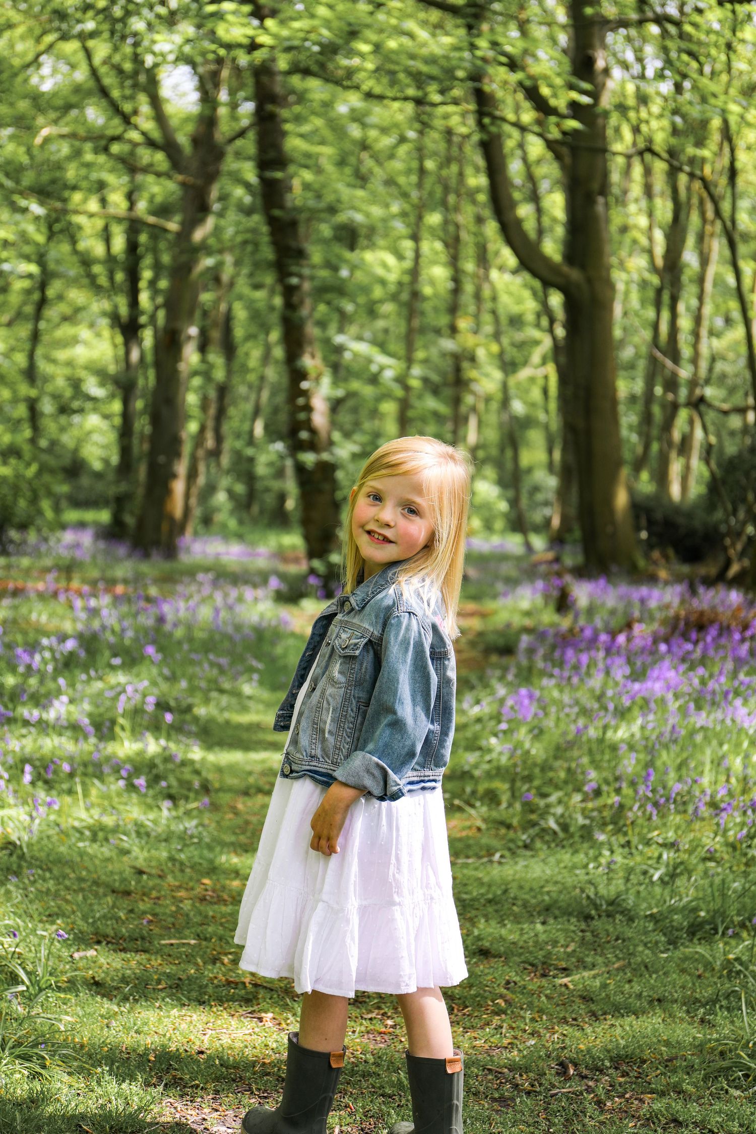 A child stands in a spring forest wearing a denim jacket and white dress among blooming purple bluebells, during family shoot in Nottingham