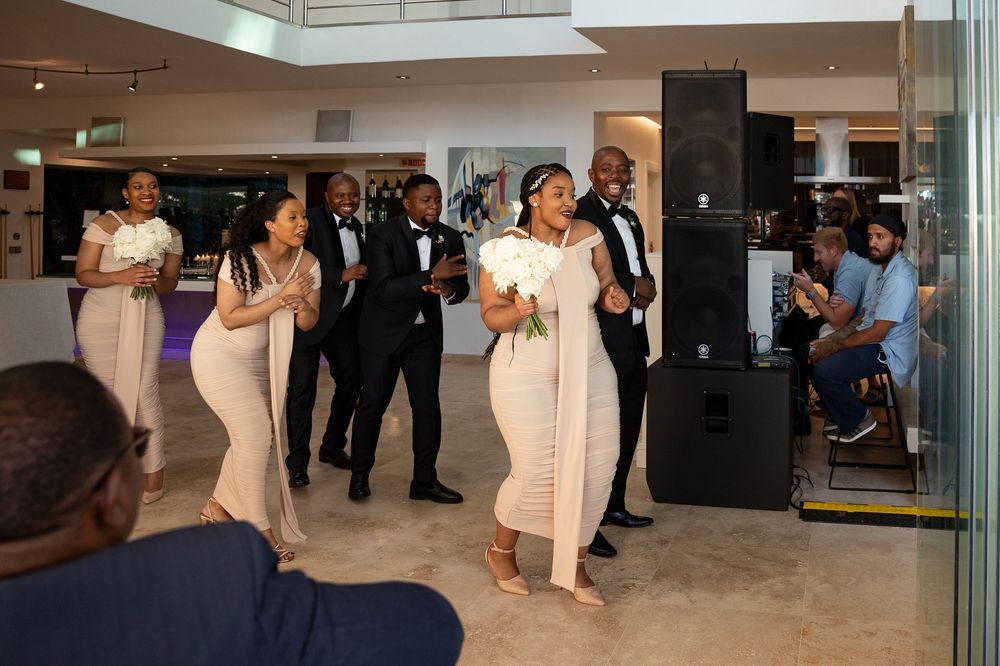 Wedding party walks and dances through a modern lobby space in coordinated pink and white attire.
