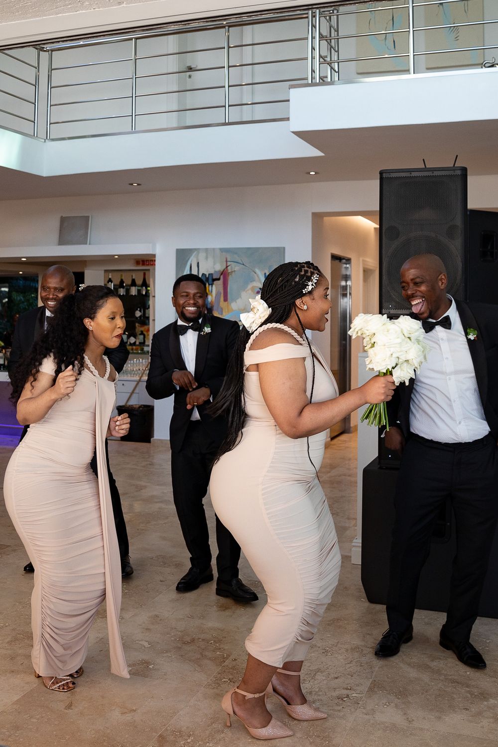 A group of wedding guests in formal attire dancing and laughing together at an indoor reception venue.
