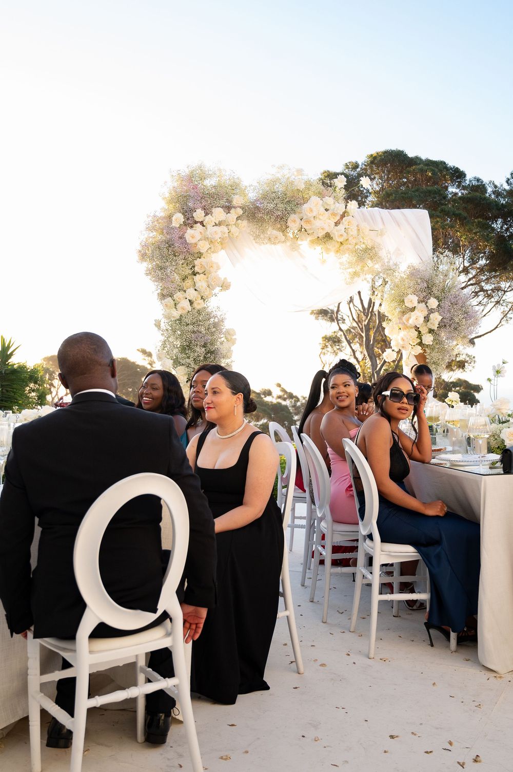 Wedding guests sit and mingle at outdoor reception tables decorated with white chairs.