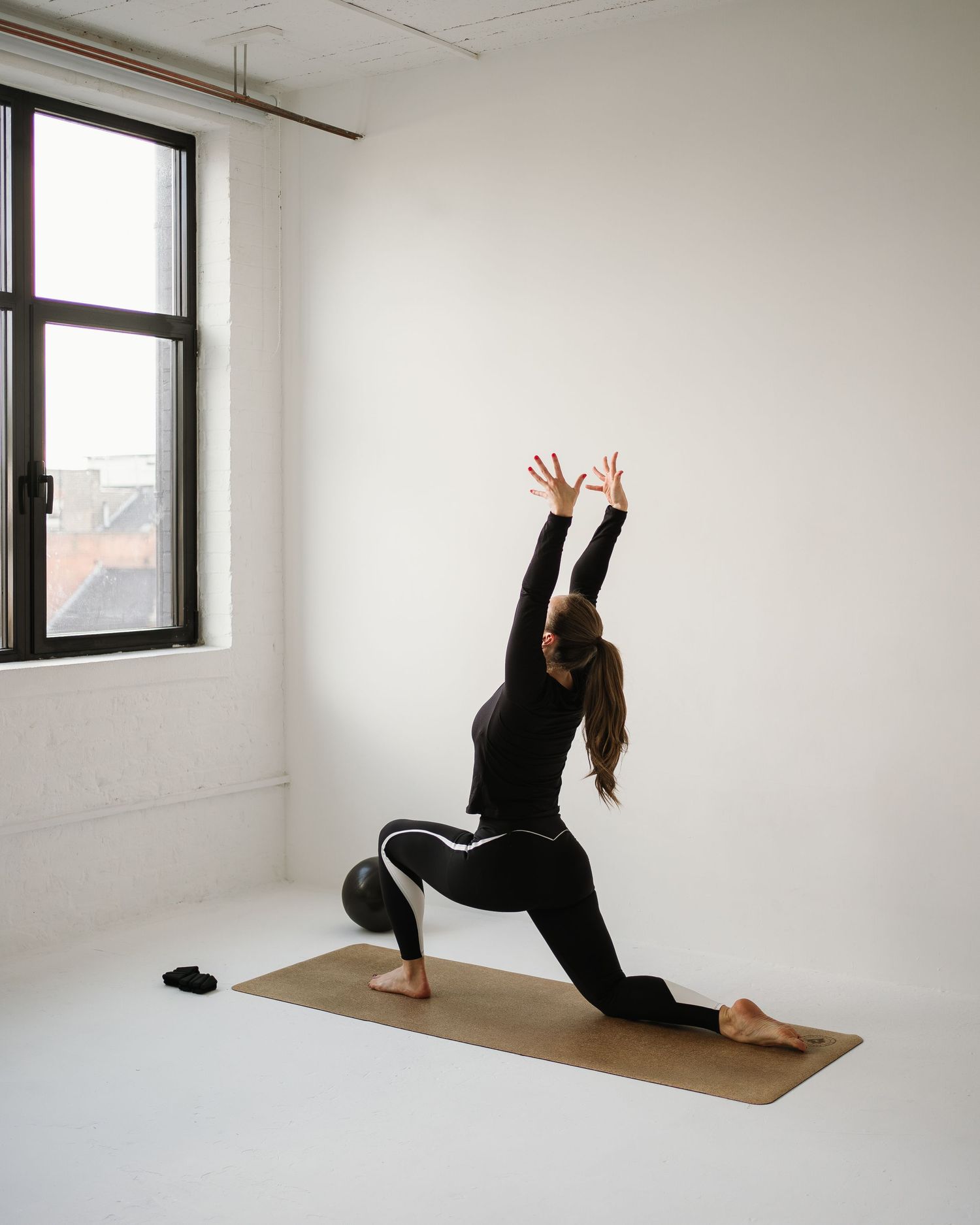 Person in black athletic wear practicing a crescent lunge yoga pose on brown mat by window in minimalist white room.