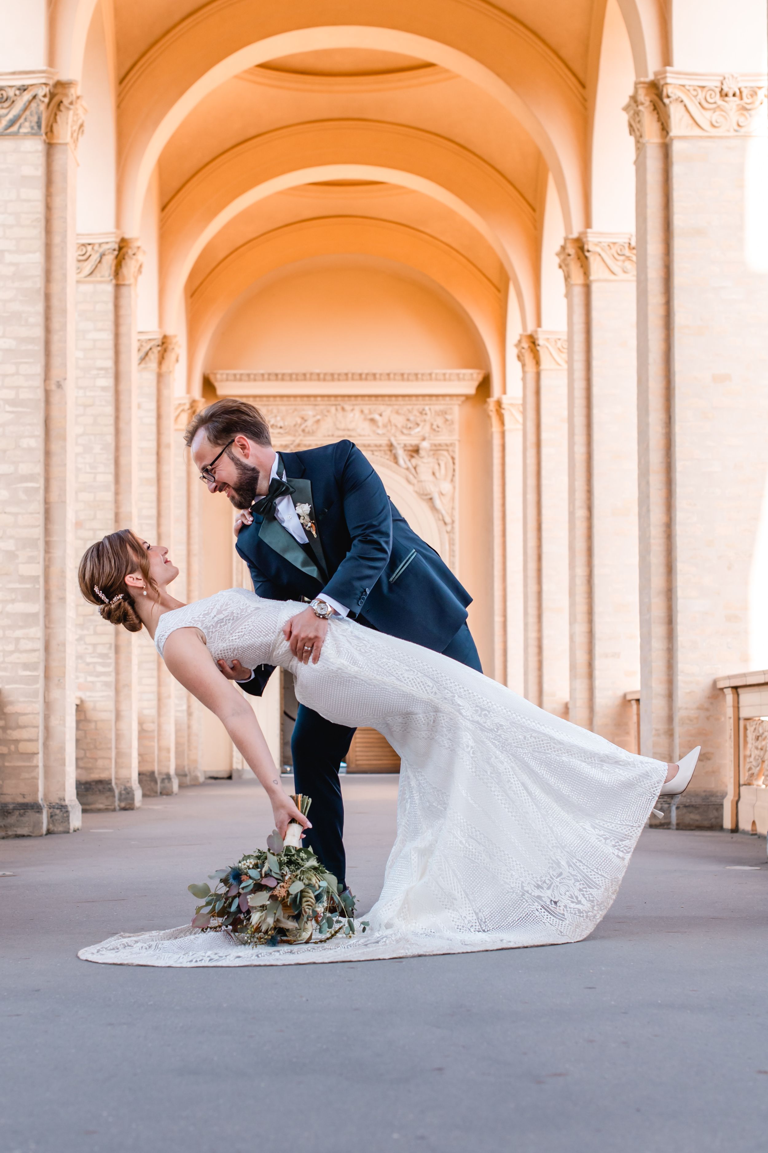 A couple in wedding attire share a romantic dance pose under ornate arched columns at sunset.