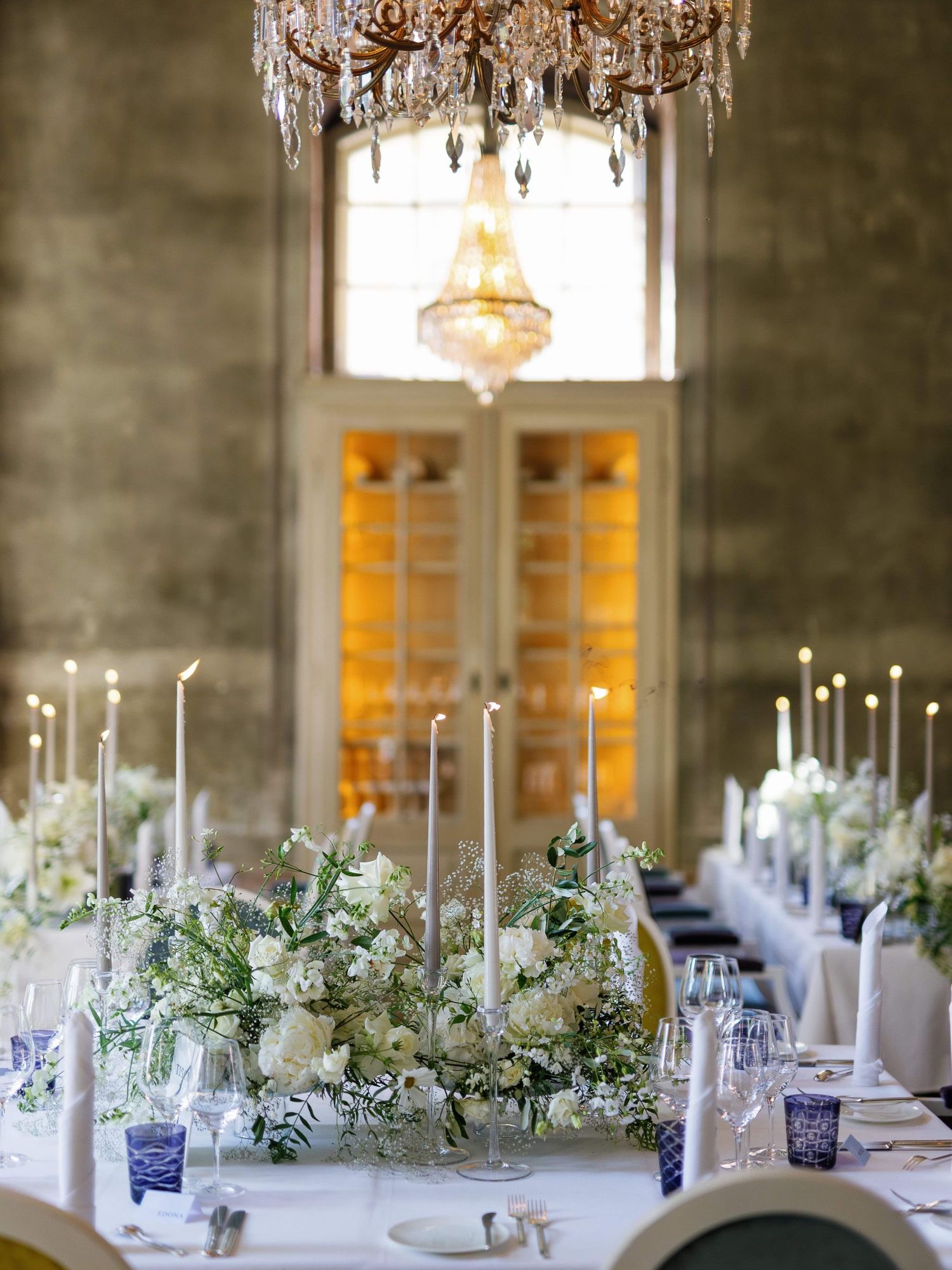 An elegant wedding reception table with white floral centerpieces, tall candles, and a crystal chandelier overhead.