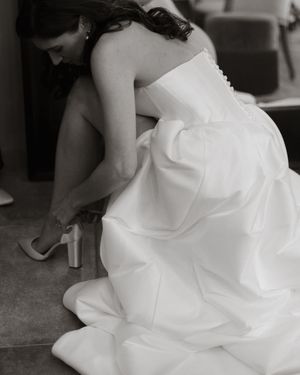 A bride in a white wedding dress adjusts her silver high heel shoe while seated in a black and white photograph.