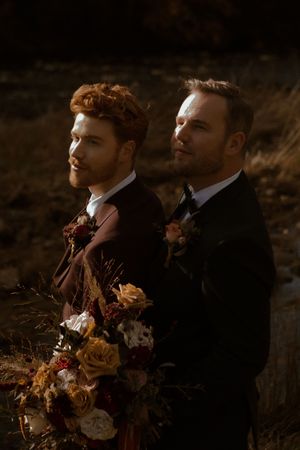 Two people in formal suits stand together outdoors holding autumn-colored floral bouquets in moody lighting.